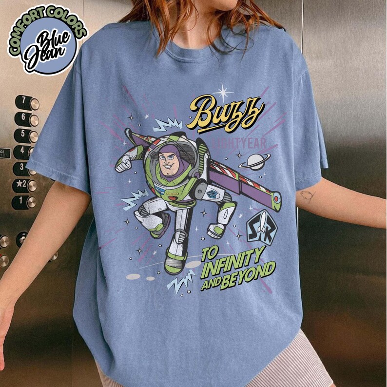 May include: Blue t-shirt featuring a graphic of Buzz Lightyear with the text "Buzz Lightyear" and "To Infinity and Beyond." The shirt has a round Comfort Colors logo in the upper left corner.