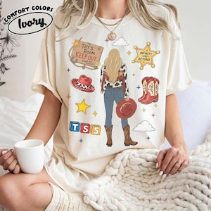 May include: An ivory short-sleeve t-shirt featuring a graphic print of a person with long blonde hair, wearing a cow-print jacket, blue jeans, and brown boots. The design includes a wooden sign reading Tay's Room Keep Out, a sheriff star, red cowboy hats, and blocks labelled TS5.