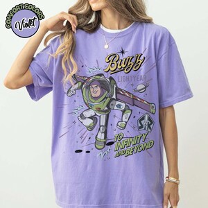 May include: A violet Comfort Colors t-shirt featuring a graphic of Buzz Lightyear in his spacesuit. The design includes the text "Buzz Lightyear" and "To Infinity and Beyond" in a comic book style. The shirt has a crew neck.
