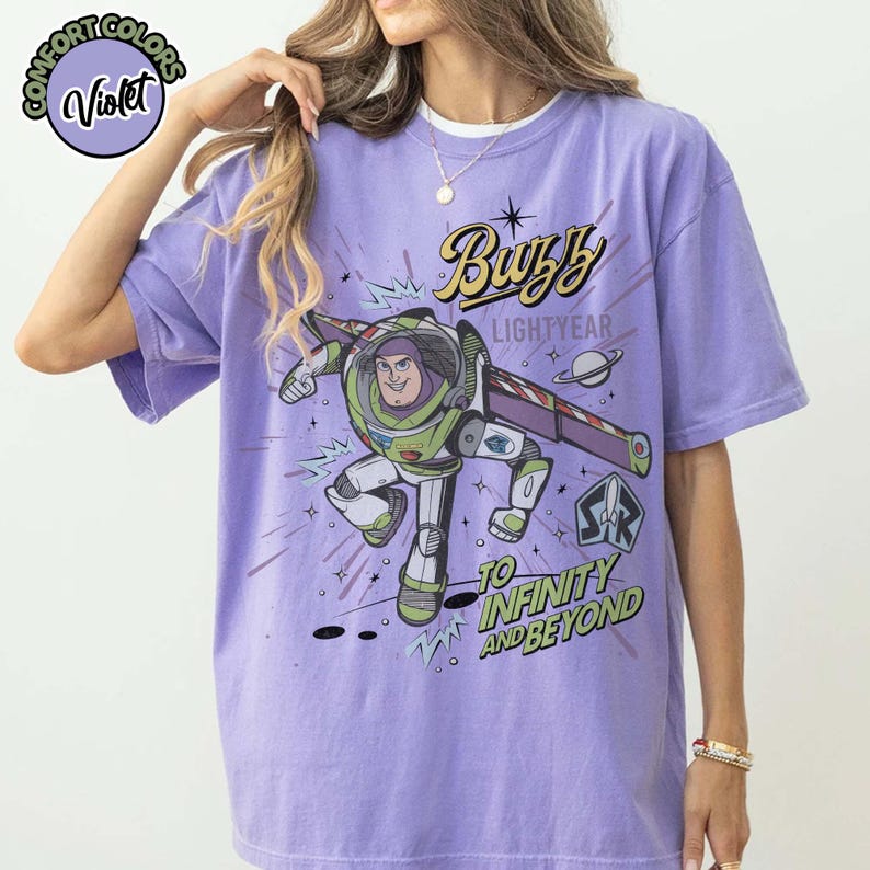 May include: A violet Comfort Colors t-shirt featuring a graphic of Buzz Lightyear in his spacesuit. The design includes the text "Buzz Lightyear" and "To Infinity and Beyond" in a comic book style. The shirt has a crew neck.