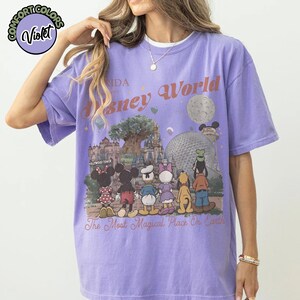 May include: A violet-colored t-shirt featuring a graphic of Disney World with various characters and park landmarks. The text "Disney World" and "The Most Magical Place On Earth" are printed on the front. The shirt has a crew neck.