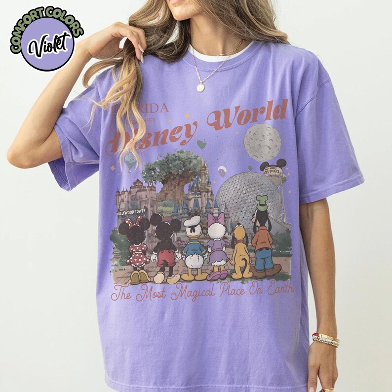 May include: A violet-colored t-shirt featuring a graphic of Disney World with various characters and park landmarks. The text "Disney World" and "The Most Magical Place On Earth" are printed on the front. The shirt has a crew neck.