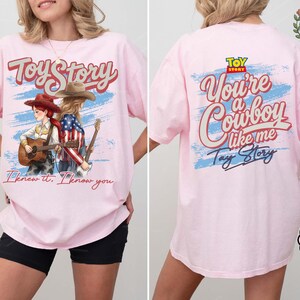 May include: A light pink t-shirt featuring a front graphic of two figures in cowboy hats playing guitars with the text Toy Story and I knew it, I know you. The back displays the text Toy Story, You're a Cowboy like me, and Tay Story.