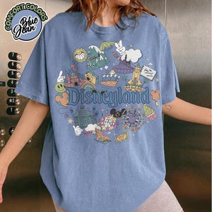Puede incluir: Camiseta azul vaquero con un gr&aacute;fico de Disneyland. El gr&aacute;fico presenta varios iconos de Disneyland, incluyendo un castillo, una noria y Mickey Mouse. La camiseta tiene cuello redondo y mangas cortas. Las palabras "Comfort Colors" y "Blue Jean" est&aacute;n impresas en la parte superior izquierda.