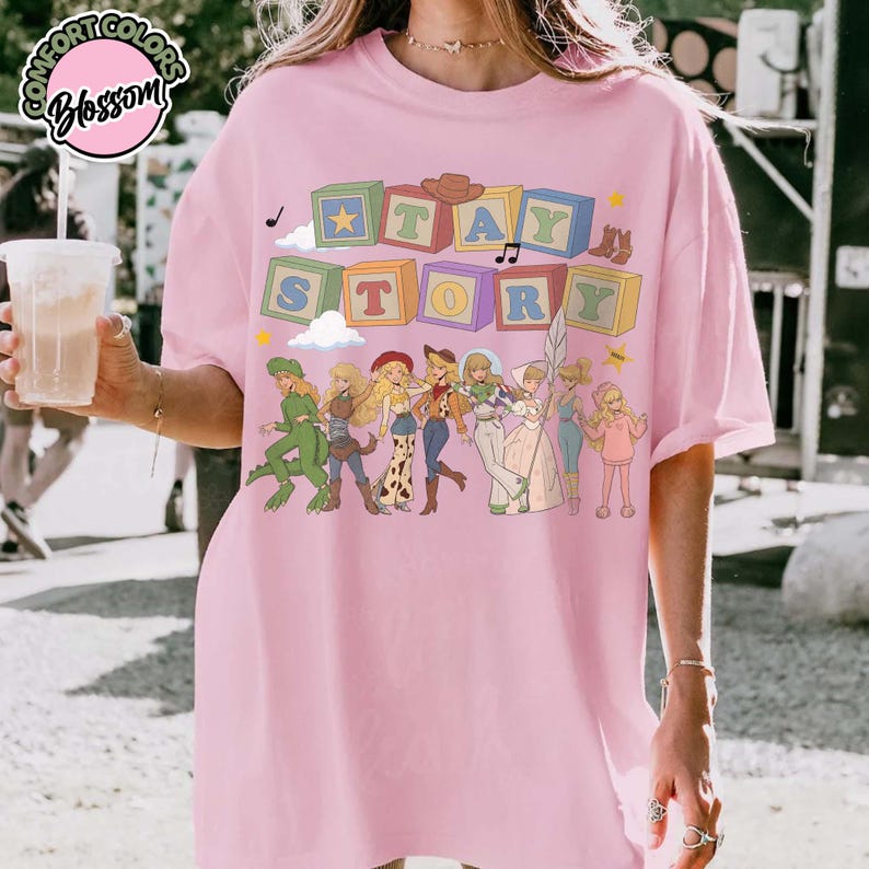 May include: A light pink t-shirt featuring a graphic print of colourful alphabet blocks spelling the word STAY STORY above a line of illustrated female characters in various outfits, including a green dinosaur costume and western attire.