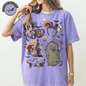 May include: Violet Comfort Colors t-shirt featuring a colorful graphic design with characters and elements from "The Nightmare Before Christmas." The design includes Jack Skellington, Sally, Oogie Boogie, and other iconic imagery. The text "NIGHTMARE" is also visible.