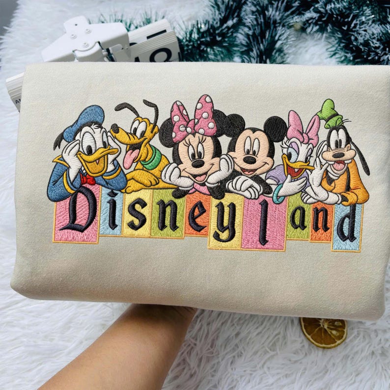 May include: A beige sweatshirt featuring an embroidered graphic of Donald Duck, Pluto, Minnie Mouse, Mickey Mouse, Daisy Duck, and Goofy. Below the characters, the word Disneyland is embroidered in black letters over colorful rectangular blocks.