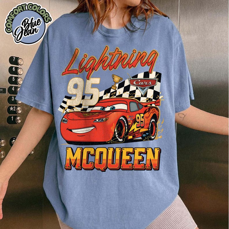 Pu&ograve; includere: T-shirt azzurra con grafica di Saetta McQueen del film Cars. Il design include la scritta "Lightning 95 McQueen" con una bandiera a scacchi e un trofeo. La maglietta ha un aspetto vintage e usurato.
