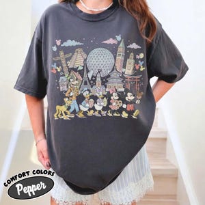 May include: Dark gray t-shirt featuring a colorful graphic of Disney characters and world landmarks. The design includes Mickey Mouse, Donald Duck, and other characters. The shirt has the text "COMFORT COLORS Pepper" at the bottom.
