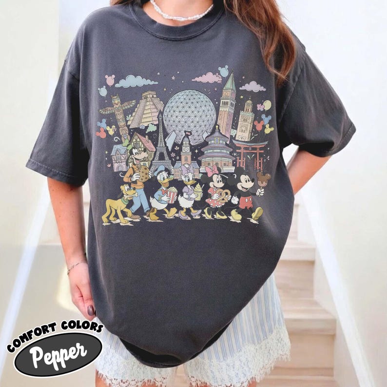 May include: Dark gray t-shirt featuring a colorful graphic of Disney characters and world landmarks. The design includes Mickey Mouse, Donald Duck, and other characters. The shirt has the text "COMFORT COLORS Pepper" at the bottom.