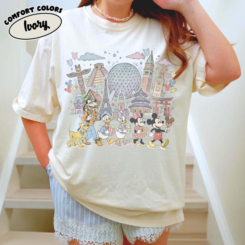 May include: Ivory-colored t-shirt with a colorful graphic featuring cartoon characters and landmarks. The graphic includes Mickey Mouse, Donald Duck, and other characters, along with the Eiffel Tower and other world landmarks. The text "COMFORT COLORS" and "Ivory" are printed on the shirt.