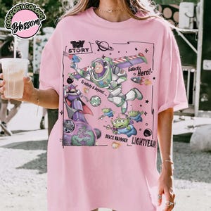 Puede incluir: Una camiseta rosa claro con un estampado gr&aacute;fico de Buzz Lightyear, Zurg y alien&iacute;genas verdes de Toy Story. El dise&ntilde;o incluye el texto "Toy Story", "Galactic Hero!", "To infinity & beyond...", "Space Ranger" y "Lightyear" en tinta negra.