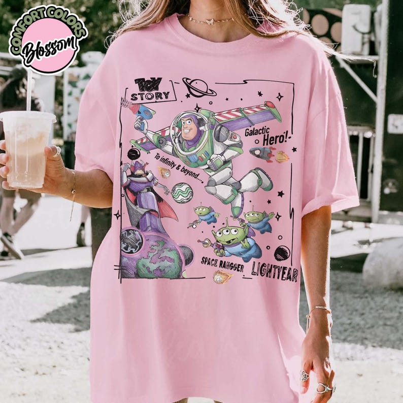 Puede incluir: Una camiseta rosa claro con un estampado gr&aacute;fico de Buzz Lightyear, Zurg y alien&iacute;genas verdes de Toy Story. El dise&ntilde;o incluye el texto "Toy Story", "Galactic Hero!", "To infinity & beyond...", "Space Ranger" y "Lightyear" en tinta negra.