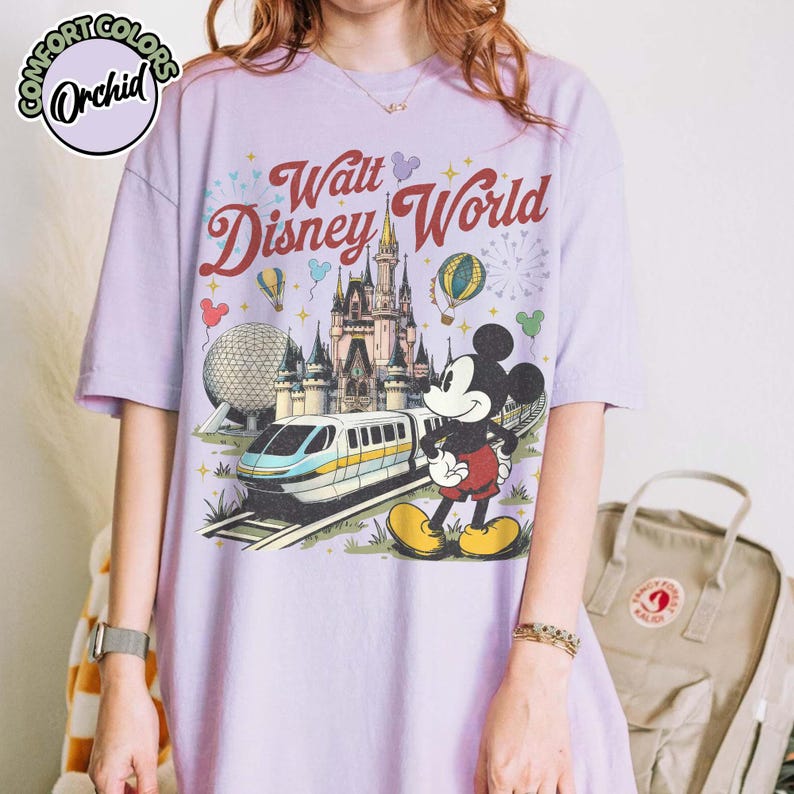 May include: A lilac t-shirt with a graphic of Mickey Mouse and Cinderella's castle at Walt Disney World. The design includes a monorail, Spaceship Earth, and hot air balloons. The text "Walt Disney World" is in a retro style.