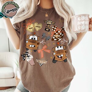 Puede incluir: Camiseta marr&oacute;n con im&aacute;genes de dibujos animados de Mate del coche de la pel&iacute;cula Cars, junto con otros gr&aacute;ficos como corazones, lazos y un trofeo. La camiseta tiene la palabra "Cars" impresa.