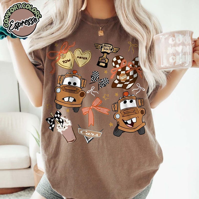 Puede incluir: Camiseta marr&oacute;n con im&aacute;genes de dibujos animados de Mate del coche de la pel&iacute;cula Cars, junto con otros gr&aacute;ficos como corazones, lazos y un trofeo. La camiseta tiene la palabra "Cars" impresa.