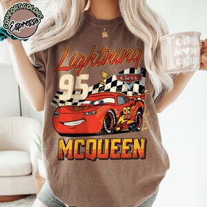 Pu&ograve; includere: T-shirt marrone con una grafica di Saetta McQueen dal film Cars. Il design include il testo "Lightning 95 McQueen" in stile vintage, con una bandiera a scacchi e un trofeo.