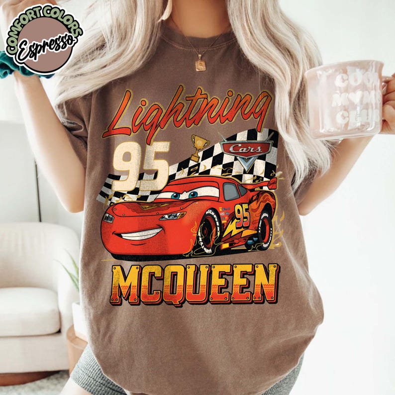 Pu&ograve; includere: T-shirt marrone con una grafica di Saetta McQueen dal film Cars. Il design include il testo "Lightning 95 McQueen" in stile vintage, con una bandiera a scacchi e un trofeo.