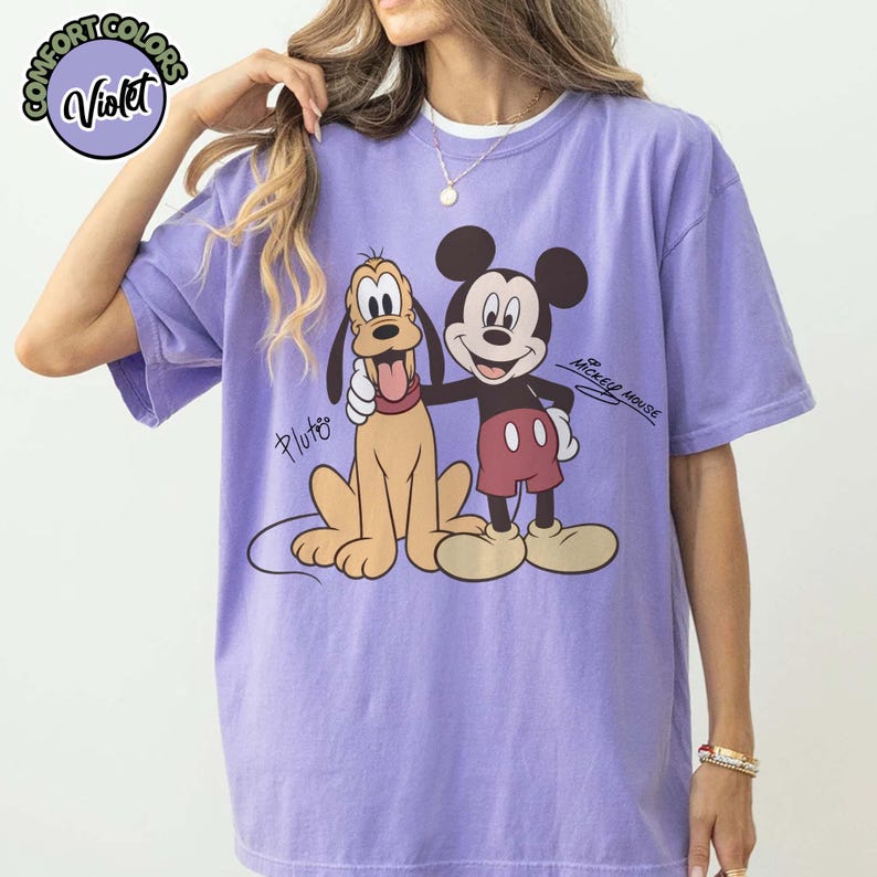 May include: Violet Comfort Colors t-shirt featuring Mickey Mouse and Pluto. Mickey Mouse is wearing red shorts and Pluto is tan. The shirt has the words "Mickey Mouse" and "Pluto" printed on it.