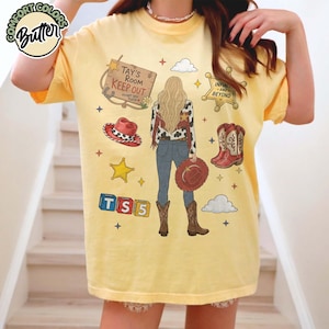 May include: A pale yellow t-shirt featuring a graphic of a person with long blonde hair seen from behind, wearing a cow-print jacket, blue jeans, and brown boots. The design includes a sign reading Tay's Room Keep Out, a sheriff badge, cowboy hat, boots, and TS5 blocks.