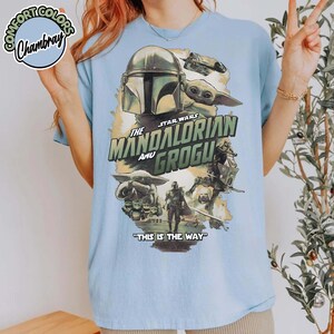 May include: A light blue t-shirt featuring a graphic print of The Mandalorian and Grogu. The design includes the characters, a spaceship, and the text "The Mandalorian and Grogu" along with the phrase "This is the way" printed in dark green.