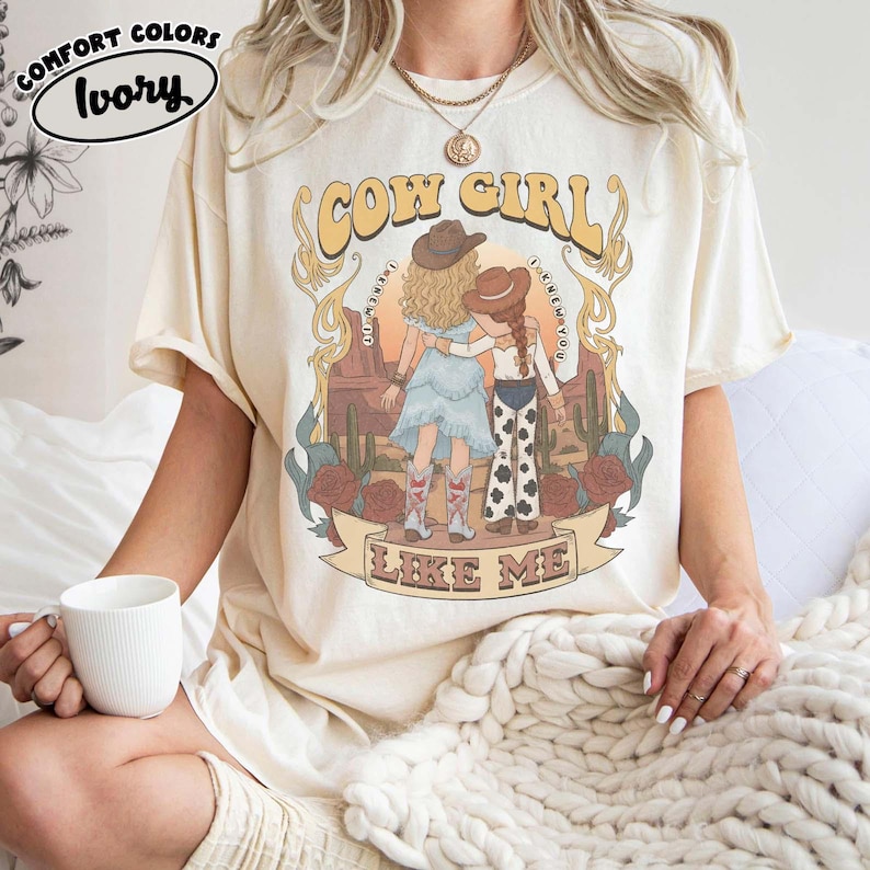 May include: An ivory short-sleeve t-shirt featuring a graphic print of two figures in cowboy hats standing in a desert landscape with cacti and roses. The text reads COW GIRL LIKE ME. A Comfort Colors Ivory label is visible in the upper left corner.
