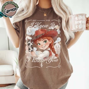 Peut inclure: Un t-shirt marron &agrave; manches courtes avec une illustration de cow-girl portant un chapeau rouge et un gant &agrave; motifs de vache. Le texte indique EST 2026, I Knew It et I Knew You, entour&eacute; d'&eacute;toiles, de fers &agrave; cheval et d'une note de musique.
