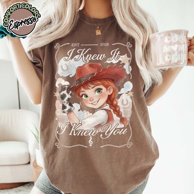 Peut inclure: Un t-shirt marron &agrave; manches courtes avec une illustration de cow-girl portant un chapeau rouge et un gant &agrave; motifs de vache. Le texte indique EST 2026, I Knew It et I Knew You, entour&eacute; d'&eacute;toiles, de fers &agrave; cheval et d'une note de musique.