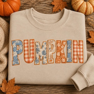 Puede incluir: Una sudadera beige de cuello redondo con la palabra PUMPKIN en el pecho. Cada letra está rellena con un patrón de tela diferente, incluyendo flores azules, cuadros vichy naranjas y blancos, y hojas naranjas, bordeado con hilo naranja.