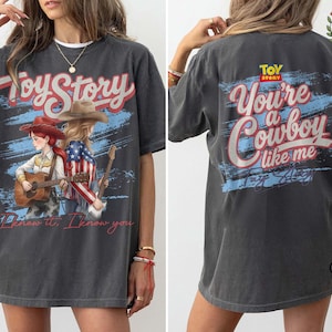 May include: A charcoal gray t-shirt featuring a Toy Story graphic. The front displays two cowgirls with guitars and the text "I knew it, I know you." The back features the Toy Story logo and the text "You're a Cowboy like me" in red and white.