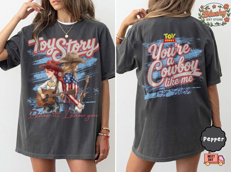 May include: A charcoal gray t-shirt featuring a Toy Story graphic. The front displays two cowgirls with guitars and the text "I knew it, I know you." The back features the Toy Story logo and the text "You're a Cowboy like me" in red and white.