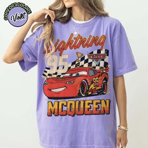 Pu&ograve; includere: Una maglietta viola con una grafica di Saetta McQueen del film Cars. Il design include il numero 95, una bandiera a scacchi e la parola "McQueen". La maglietta ha maniche corte e girocollo.