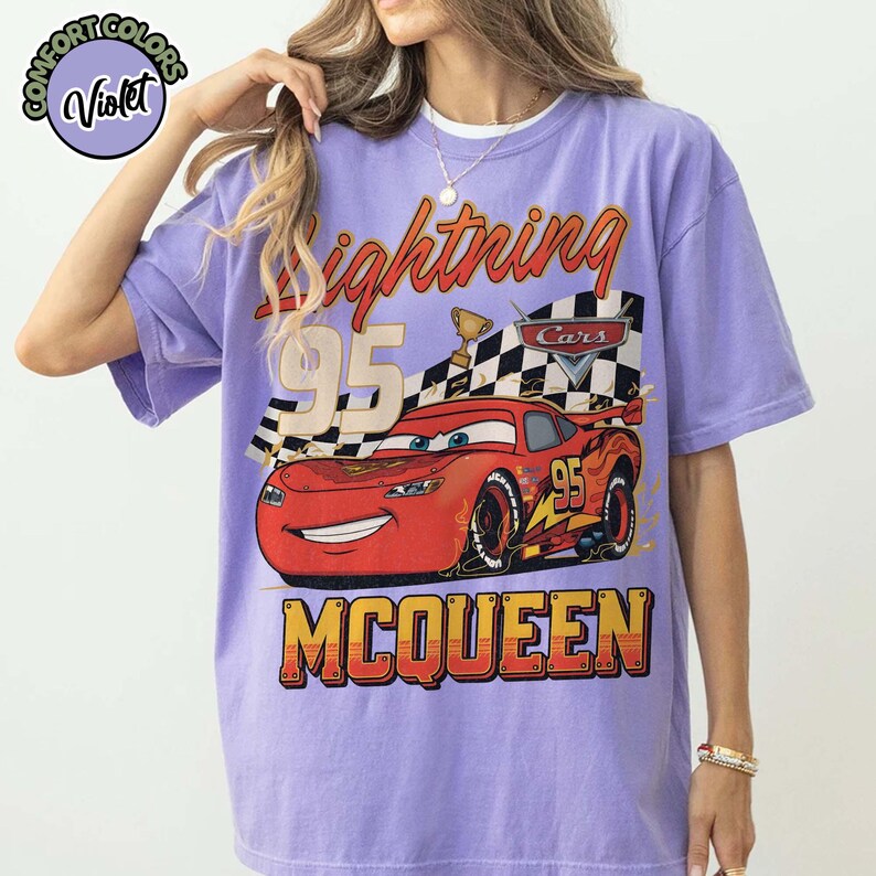 Pu&ograve; includere: Una maglietta viola con una grafica di Saetta McQueen del film Cars. Il design include il numero 95, una bandiera a scacchi e la parola "McQueen". La maglietta ha maniche corte e girocollo.