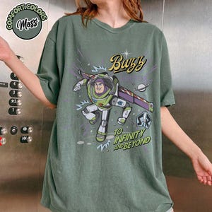 May include: Moss green t-shirt featuring a graphic of Buzz Lightyear with the text "Buzz Lightyear" and "To Infinity and Beyond". The shirt has short sleeves and a crew neck. The shirt is made of a soft, comfortable material.