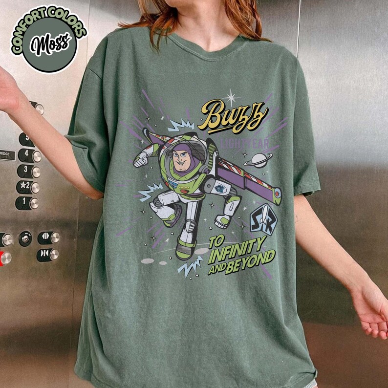 May include: Moss green t-shirt featuring a graphic of Buzz Lightyear with the text "Buzz Lightyear" and "To Infinity and Beyond". The shirt has short sleeves and a crew neck. The shirt is made of a soft, comfortable material.