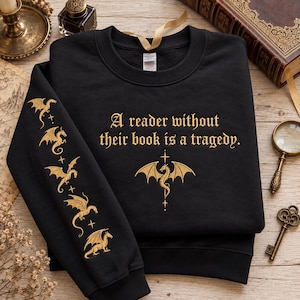 May include: A black crewneck sweatshirt featuring gold embroidery. The chest displays the text A reader without their book is a tragedy. above a dragon icon. The left sleeve features a vertical row of five small gold embroidered dragons.