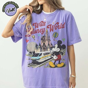 May include: A lilac t-shirt with a graphic of Mickey Mouse, a monorail, a castle, and the text "Walt Disney World." The design includes hot air balloons and the Epcot ball. The shirt has short sleeves.