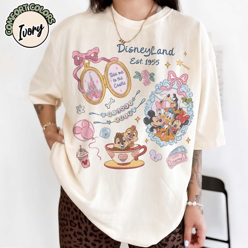 Peut inclure: T-shirt ivoire avec des graphismes de Disneyland. Le design comprend un m&eacute;daillon avec un ch&acirc;teau, Mickey Mouse et ses amis, Tic et Tac dans une tasse &agrave; th&eacute;, et le texte "Disneyland Est. 1955".