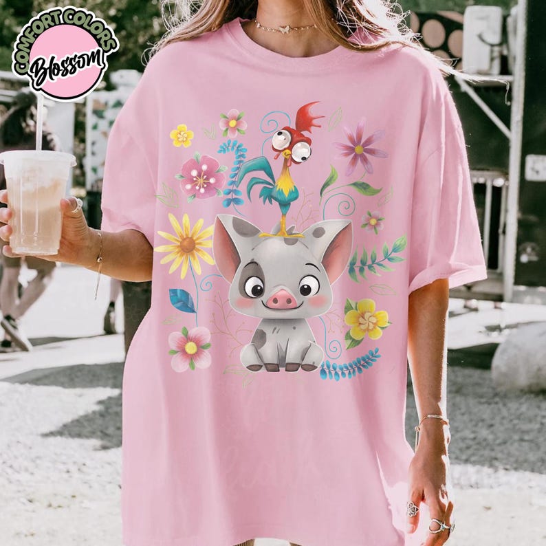 May include: Pink t-shirt featuring a cartoon pig and rooster with floral accents. The design includes a pig with gray spots and a colorful rooster. The shirt has the text "Comfort Colors Blossom" in a circular design.
