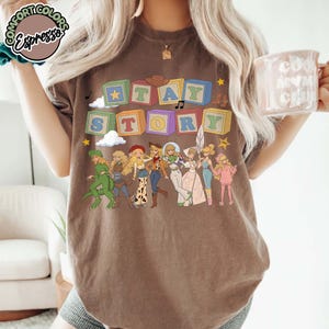 May include: A brown t-shirt featuring colourful alphabet blocks spelling out "TAY STORY" above a line of illustrated characters dressed in various outfits. The design includes small clouds, stars, and musical notes, with a "COMFORT COLORS Espresso" logo.
