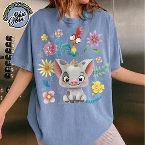 May include: Blue graphic tee featuring a cartoon pig and rooster surrounded by colorful flowers and leaves. The shirt has the text "Comfort Colors Blue Jean" in a circular design. The tee is a light blue color.