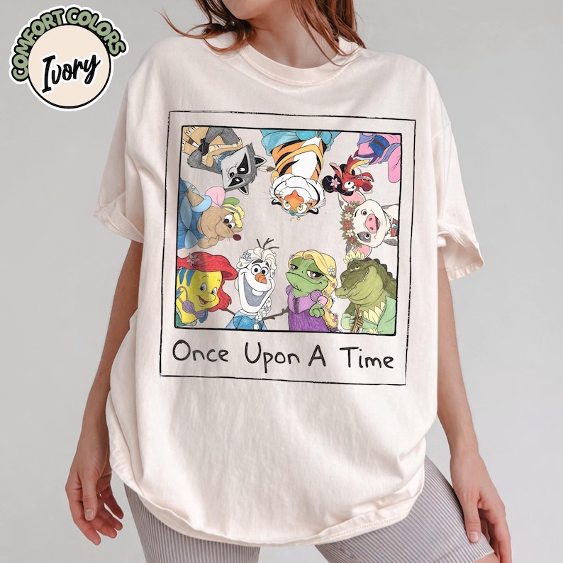May include: Ivory-colored t-shirt featuring a framed graphic of cartoon characters, including a raccoon, tiger, and frog. The graphic is labeled "Once Upon A Time". The shirt has short sleeves and a crew neck.