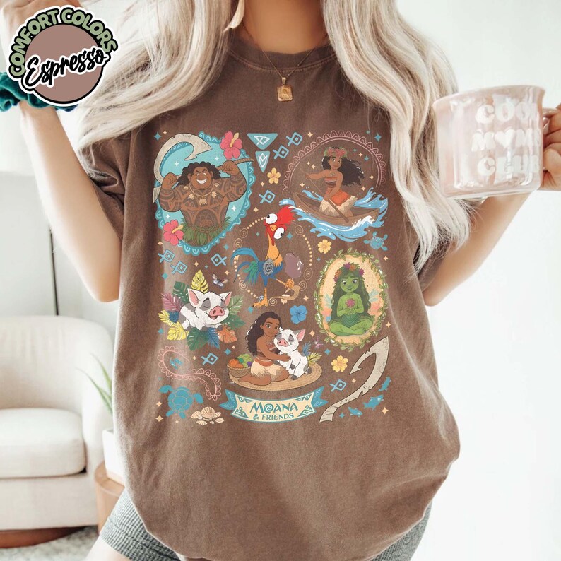 May include: A brown short-sleeve t-shirt featuring a graphic print of Moana and friends, including Maui, Pua, Heihei, and Te Fiti. The design incorporates tropical flowers, tribal patterns, and a banner with the text Moana & Friends.