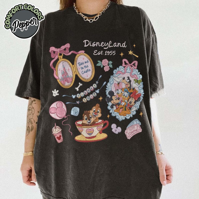 Peut inclure: T-shirt gris fonc&eacute; avec des graphismes de Disneyland. Le design comprend des personnages Disney, un ch&acirc;teau et le texte "Disneyland Est. 1955". Des graphismes suppl&eacute;mentaires incluent une tasse &agrave; th&eacute;, un ballon et un billet.