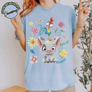 May include: Light blue t-shirt featuring a cartoon pig and rooster design with floral accents. The design includes a gray pig with pink ears and a red, yellow, and blue rooster. The shirt has the text "Comfort Colors Chambray" in a circle.