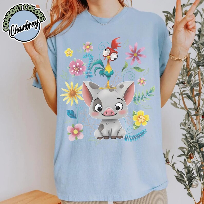 May include: Light blue t-shirt featuring a cartoon pig and rooster design with floral accents. The design includes a gray pig with pink ears and a red, yellow, and blue rooster. The shirt has the text "Comfort Colors Chambray" in a circle.