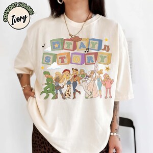 May include: An ivory t-shirt featuring a graphic print of colourful alphabet blocks spelling out TAY STORY. Below the blocks are illustrations of eight characters in various outfits, including a dinosaur suit, cowgirl attire, and space gear.