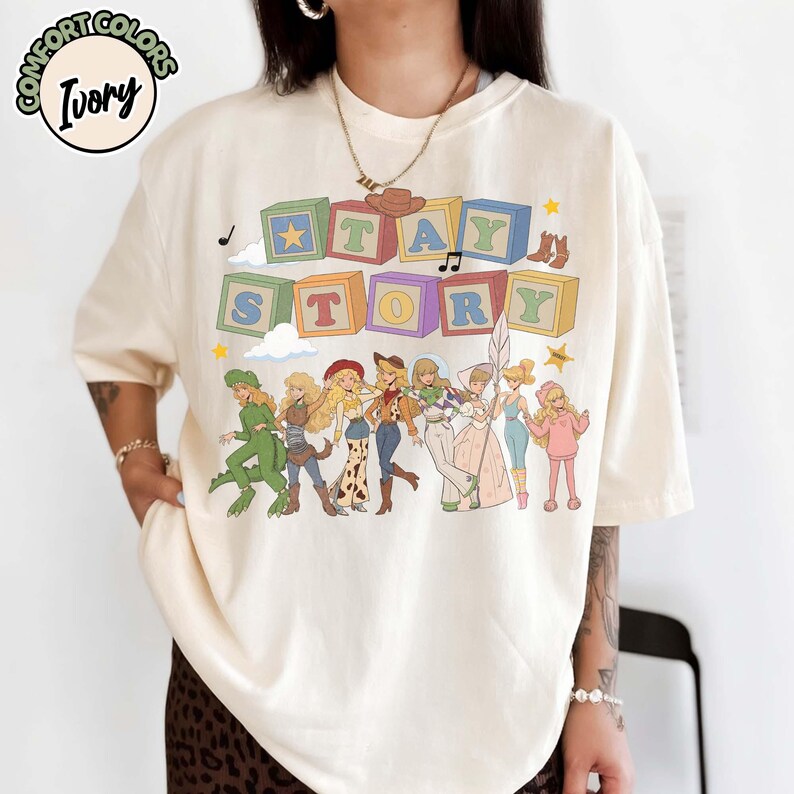 May include: An ivory t-shirt featuring a graphic print of colourful alphabet blocks spelling out TAY STORY. Below the blocks are illustrations of eight characters in various outfits, including a dinosaur suit, cowgirl attire, and space gear.