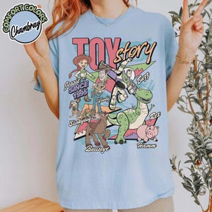 May include: Light blue t-shirt featuring the words "Toy Story" in a bold, colorful design. The shirt showcases various characters including Woody, Buzz Lightyear, Jessie, Rex, Hamm, Slinkydog, and Bullseye. The text "Since 1995" is also included.