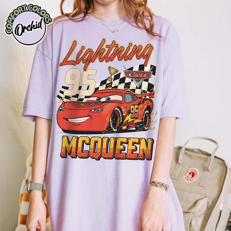 Pu&ograve; includere: Una maglietta lilla con una grafica di Saetta McQueen del film Cars. Il design include il testo "Lightning 95 McQueen" con una bandiera a scacchi e un trofeo. La maglietta ha maniche corte e girocollo.